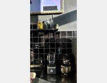 Küchenarbeitsbereich mit schwarzer glänzender Fliesenwand, einem Mixer und einer Küchenmaschine auf einer dunklen Arbeitsplatte. Darüber befindet sich ein Regal mit Gläsern, einer Vase mit Zitronen und anderen kleinen Gegenständen. Im oberen Bereich hängt ein gerahmtes Poster mit englischem Text.