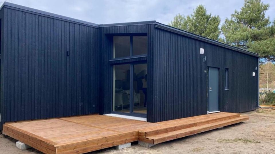 Modernes, eingeschossiges Haus mit schwarzer Holzfassade und Pultdach, davor eine große Holzterrasse mit Stufen. Mittig befinden sich bodentiefe Glasfenster, rechts eine Außentür; im Hintergrund sind Bäume und ein offener, sandiger Boden zu sehen.