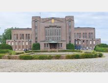 Backsteinbau im traditionellen Stil mit symmetrischer Fassade und großen Sprossenfenstern; im Vordergrund ein kreisförmiger Brunnen, umgeben von niedrigen Büschen und einem gepflasterten Platz.