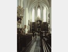 Innenansicht einer gotischen Kirche mit hohen Spitzbögen, großen Buntglasfenstern und einem reich verzierten Altar im Hintergrund. Links sind geschnitzte Statuen und ein Kanzelaufbau zu sehen, während Kirchenbänke den Gang auf beiden Seiten flankieren.