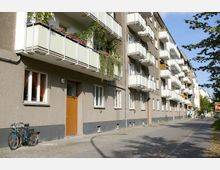 Wohnhaus im urbanen Umfeld mit brauner Putzfassade und mehreren Reihen von Balkonen mit Blumen und Pflanzen. Im Vordergrund stehen zwei Fahrräder vor einer hölzernen Eingangstür, daneben ein schmaler Gehweg.