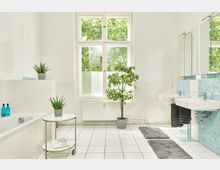 Helles Badezimmer mit weißen Fliesen und großen Fenstern, die Blick ins Grüne bieten. Ausstattung umfasst eine Badewanne, zwei Waschbecken mit blauen Mosaikfliesen, graue Teppiche, Zimmerpflanzen und ein weißer Rolltisch.