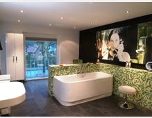 Modernes Badezimmer mit freistehender weißen Badewanne, grünen Mosaikfliesen an der Wand und dunklem Steinboden. Im Hintergrund ein großes Fenster mit Blick nach draußen und eine Wand mit einem großformatigen Kunstbild.