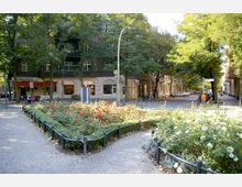 Straßenkreuzung in einem Wohngebiet mit einer bepflanzten Mittelinsel voller Rosen in Rot und Weiß, umgeben von einem niedrigen Geländer. Im Hintergrund stehen mehrstöckige Wohnhäuser mit Geschäften im Erdgeschoss, darunter eine Apotheke und ein Lokal namens „S-Bahn-Klause“.