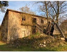 Ein verlassenes, verfallenes Gebäude aus Ziegelstein mit beschädigtem Dach und zerbrochenen Fenstern, umgeben von kahlen Bäumen und Wildwuchs. Im Vordergrund befinden sich Steine, Holzreste und Grasbewuchs.