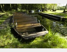 Ein flacher Holzkahn liegt an einem schmalen Steg in einem von Bäumen umgebenen, ruhigen Kanal. Im Hintergrund sind weitere Boote und eine Wiese mit Sitzbänken erkennbar.