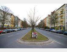 Breite, baumgesäumte Straße mit einer Grünfläche in der Mitte, gesäumt von farbigen mehrstöckigen Wohnhäusern. Parkende Autos reihen sich entlang der beiden Fahrspuren, ein Plakat ist auf der Grünfläche sichtbar.