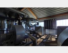 Industriehalle mit einer Anlage zur Luft- oder Ablufttechnik, bestehend aus großen Metallrohren, Lüftungskästen und technischen Komponenten. Die Halle hat ein Dach aus Wellblech und große Fenster, die Tageslicht einlassen.
