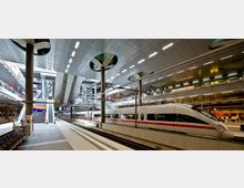 Moderner Bahnhof mit mehreren Ebenen und einem ICE-Zug auf einem der Gleise. Die Architektur umfasst breite Treppen, Glasgeländer und eine helle Decke mit eingebauten Lichtpaneelen, umgeben von Schildern, Geschäften und Sitzbereichen.