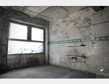 Verlassener, stark beschädigter Innenraum mit abblätterndem Putz, gemauerter Tonnenwölbung und großem, milchigem Fenster an der linken Wand. An der rechten Wand befinden sich weiße Wandfliesen mit blaugrünem Streifen sowie ein kleines Waschbecken mit Ablage.