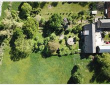 Luftaufnahme eines grünen Grundstücks mit Wiesen, Bäumen und einem Gebäudeensemble, bestehend aus Wohn- und Nebengebäuden mit Solarpanelen auf dem Dach. Im Gartenbereich sind einzelne baumbestandene Flächen, ein rundes Pavillon-Dach und ein kleiner Weg sichtbar.