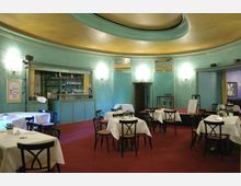 Kleiner Saal eines Restaurants oder Cafés mit türkisen Wänden, einer runden Decke mit goldfarbenem Element und rotem Teppichboden. Mehrere Tische mit weißen Tischdecken und schwarzen Stühlen sind verteilt, im Hintergrund befindet sich eine Bar mit Gläsern und einer Kaffeemaschine.