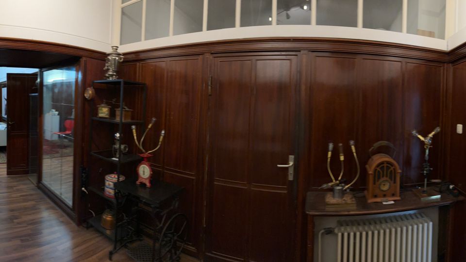 Innenansicht eines historischen Museums- oder Ausstellungsraums mit dunkler Holzvertäfelung, Parkettboden und Vitrinen sowie alten technischen Geräten und Messinstrumenten auf Tischen. Mehrere Türen und ein verglaster Durchgang verbinden den Raum mit angrenzenden Bereichen.