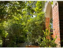 Ein kleiner Garten mit dichtem, grünem Laub und Schatten spendenden Bäumen. Im Vordergrund ist eine Backsteinmauer mit einem Rundbogen sichtbar, der den Eingang zu einer Terrasse oder einem überdachten Bereich markiert.