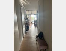 Flur eines modernen Wohnbereichs mit Holzboden, einer Bank mit einer Tasche darauf rechts und dekorativen Elementen links. Im Hintergrund sind Glastüren und große Fenster zu erkennen, die Tageslicht hereinlassen und einen Blick auf einen Außenbereich bieten.