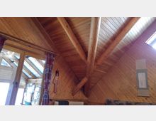 Holzvertäfeltes Dachzimmer mit sichtbaren Holzträgern und schrägen Decken. Links befindet sich eine große Glastür mit Vorhängen, die zu einem verglasten Anbau mit Dachfenstern führt.