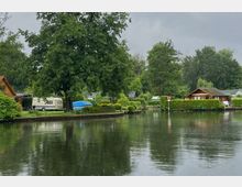 Ein ruhiger Bereich an einem Kanal mit Wasser im Vordergrund, umgeben von Bäumen, Wohnwagen, kleinen Holzhäusern und gepflegten Gärten. Im Hintergrund sind ein bewölkter Himmel, Büsche und weitere Vegetation zu sehen.