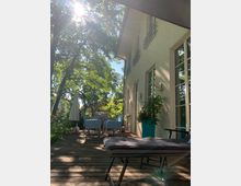 Sonnendurchflutete Holzterrasse mit Gartenmöbeln vor einer hellen Hausfassade mit großen Fenstern; im Hintergrund stehen Bäume, und links ist eine Sonnenschirmstruktur erkennbar.