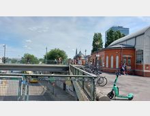 Eine Straße mit einer erhöhten Fußgängerbrücke, unter der ein Bus fährt. Rechts im Bild befindet sich ein Backsteingebäude mit Industriefenstern und Zierelementen, daneben stehen Fahrräder sowie ein E-Scooter. Im Hintergrund sind Bäume, ein Kirchturm und ein modernes Bürogebäude sichtbar.