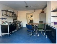 Industrieküche mit blauem Boden und weißen Wänden. Eine Arbeitsfläche mit schwarzer Metallfront, offene Regale, ein Tisch aus Holz mit verschiedenen Stühlen, ein großer Kühlschrank und eine Tür im Hintergrund definieren den Raum.