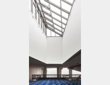 Großer, moderner Innenraum mit hohen, weißen Wänden und einem geneigten Glasdach, das viel Tageslicht einlässt. Der Boden ist mit einem blauen, gemusterten Teppich ausgelegt, und ein Geländer aus Metall umgibt eine offene Galerieebene.