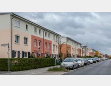 Reihenhaus-Siedlung mit hellen, pastellfarbenen Fassaden in Beige, Orange und Rot, flankiert von einer schmalen Hecke. Vor den Häusern sind parkende Autos entlang einer Straße mit Gehweg zu sehen, unter einem bewölkten Himmel.