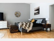 Schlafzimmer mit modernem Design: Ein graues Polsterbett mit mehreren Kissen und einer fellartigen Decke steht auf hellem Holzfußboden. Daneben befinden sich Nachttische mit Lampen sowie eine Kommode. Im Hintergrund sind Dekorationen an der grauweißen Wand, darunter ein Metall-Wandrelief und ein gerahmtes Foto. Auf dem Boden steht ein alter, abgenutzter Koffer.