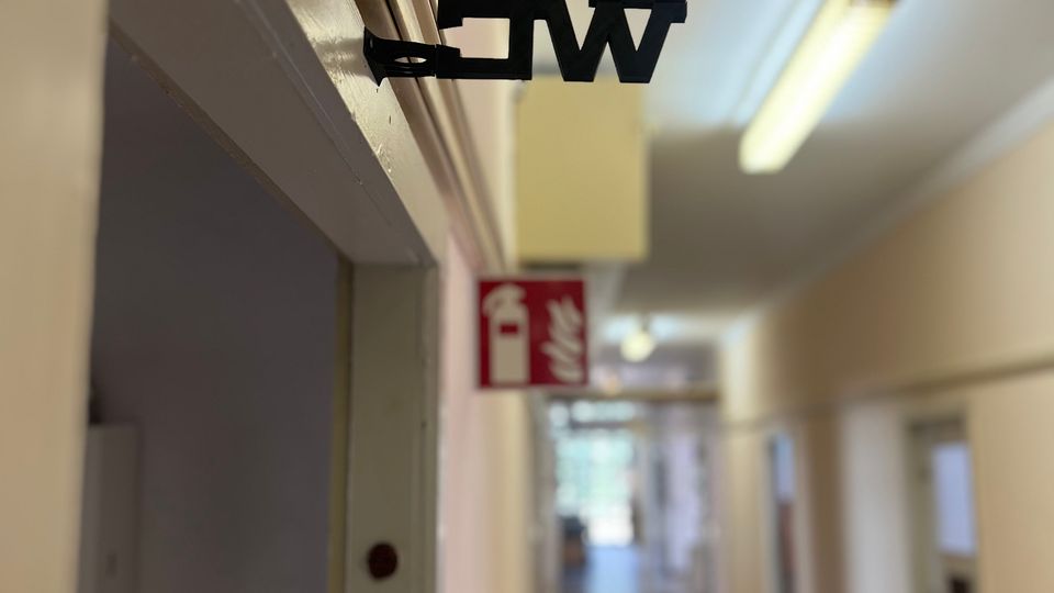 Langer Innenflur eines Gebäudes mit mehreren Türen entlang der Wände, grauem Boden und Deckenleuchten. Im Vordergrund hängt über einem Türrahmen ein schwarzes „WC“-Schild; weiter hinten ist ein rotes Brandschutzschild zu sehen.