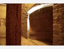 Dunkler Kellerraum mit einer massiven hölzernen Tür in einem gemauerten Rundbogen. Die Wände bestehen aus groben Ziegelsteinen, und der Boden ist aus altem Holzplanken.