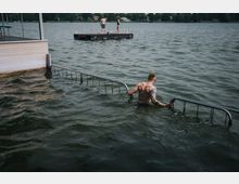 Ein Mann steht im Wasser neben einem Metallgeländer, das von einem hölzernen Steg ins Wasser führt. Im Hintergrund ist ein See mit einer kleinen, schwimmenden Plattform zu sehen, auf der zwei Personen stehen; am Ufer sind Bäume und Häuser sichtbar.