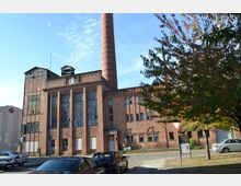 Backstein-Gebäude einer ehemaligen Fabrik mit einem hohen Schornstein, großen Fenstern und geometrischen Verzierungen an der Fassade. Im Vordergrund befinden sich parkende Autos und ein Gehweg mit Bäumen, die teilweise Schatten spenden.