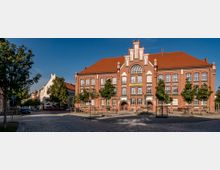 Historisches Gebäude aus rotem Backstein mit großen Fenstern, verziert mit weißen Akzenten und einem steilen roten Dach, umgeben von gepflasterten Straßen und kleinen Bäumen. Im Vordergrund steht ein Brunnen in einer offenen Platzfläche.