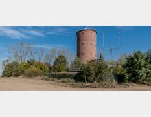 Roter Backsteinturm auf ländlichem Grundstück, umgeben von Bäumen und Sträuchern, mit mehreren Antennen und einer Parabolantenne im Vordergrund. Der blaue Himmel und das Gelände aus Erde und Pflanzen prägen die Umgebung.