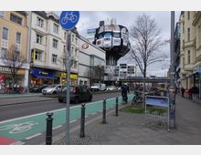 Straßenszene mit gemischter Nutzung: links moderne Geschäfte, darunter Tchibo und C&A, sowie ein Fahrradweg und parkende Fahrräder. Im Hintergrund ein markantes, futuristisches Gebäude mit aufgemalten Autos und verschiedene Schilder, die auf Einkaufsmöglichkeiten wie Saturn und Edeka hinweisen.