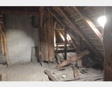 Ein verstaubter Dachboden mit sichtbaren Holzbalken, einer unvollständigen Dielenbodenstruktur und losem Schutt. Das schräge Dach zeigt freiliegende Ziegel mit einigen Lichtöffnungen, die das Innere mäßig beleuchten.