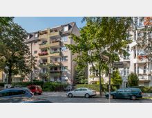 Mehrstöckiges Wohngebäude mit hellen Fassaden und Balkonen, die teilweise mit Pflanzen geschmückt sind. Im Vordergrund befindet sich eine ruhige Straße mit geparkten Autos und Straßenschildern, umgeben von Bäumen und einem kleinen Vorgarten.