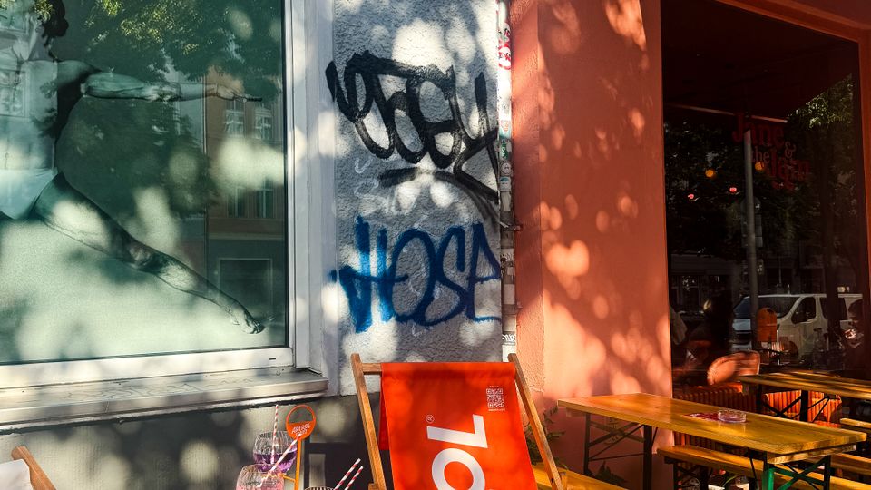 Street-side café exterior with an orange entrance wall and awning, a hanging sign reading “Jane and Jam,” and outdoor seating including wooden benches and branded deck chairs. A gray stucco wall with graffiti and a large window display sits beside small tables set with drinks, under dappled tree shade.