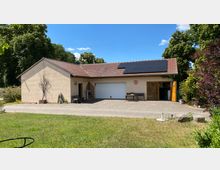 Einfamilienhaus mit hellbeigem Putz, rotem Ziegeldach und einer Photovoltaikanlage auf dem Dach. Vor dem Gebäude befinden sich eine gepflasterte Auffahrt, eine Garage mit weißem Tor sowie grüne Vegetation im Hintergrund.