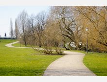 Ein Park mit einer geschwungenen, gepflasterten Gehwegführung, flankiert von grasbewachsenen Flächen und kahlen Bäumen, teilweise mit Straßenlaternen versehen. Im Hintergrund sind weitere Büsche, Bäume und ein kleiner Teich zu erkennen.