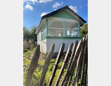 Eine kleine Gartendatsche mit weißen Wänden und türkisfarbenen Akzenten, versehen mit großen Fenstern, steht hinter einem schrägen Holzzaun in einer grünen Umgebung. Im Hintergrund sind Bäume und ein blauer Himmel mit vereinzelten Wolken zu sehen.