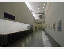 Langer, moderner Innenraum eines öffentlichen Gebäudes mit Betonwänden, einer Garderobe auf der linken Seite und einer Reihe von Plakaten an der rechten Wand. Die Decke verfügt über eine Oberlichterreihe und Einbauleuchten; am Ende des Raums befinden sich Türen.