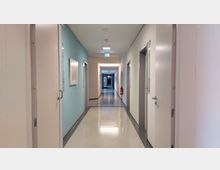 Langer, heller Flur in einem modernen Gebäude mit glatten, hellen Wänden, Türen auf beiden Seiten und einem blauen Abschnitt an der linken Wand. Es hängen vereinzelte Bilder an den Wänden, und eine Notausgangsbeleuchtung sowie ein Feuerlöscher sind zu sehen.