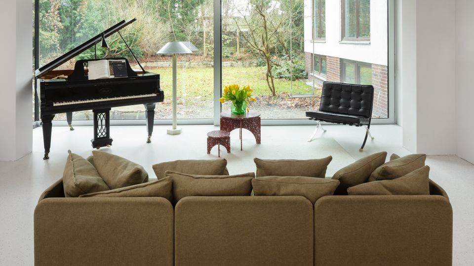 Minimalist living room with a grand piano, floor lamp, and low tables in front of a large wall of sliding glass windows overlooking a garden. A sectional sofa faces the windows, with a black leather lounge chair near the glass.