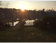 Eine Uferlandschaft bei Sonnenuntergang mit Blick auf einen ruhigen See, gesäumt von Bäumen und vereinzelten Häusern. Im Vordergrund führen eine Wiese und ein kleiner Steg mit Holzzäunen zum Wasser.