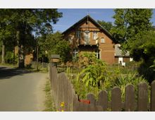 Backsteinhaus im ländlichen Stil mit Spitzdach und dekorativen Fensterumrandungen, umgeben von einem Garten mit Blumen und Pflanzen. Im Vordergrund steht ein hölzerner Zaun, und links führt eine schmale Straße entlang.