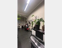 Ein moderner Tattoo-Studio-Raum mit dunklem Fliesenboden, einer Liege mit schwarzer Schutzfolie und roten sowie schwarzen Stühlen. An den Wänden hängen gerahmte Bilder, und auf einer Kommode steht eine große grüne Pflanze sowie eine Buddha-Statue.