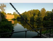 Ein ruhiger Fluss mit klarem Wasser, umgeben von dichten Bäumen und Vegetation. Im Hintergrund ist ein ziegelroter Gebäudeteil sichtbar, während im Vordergrund eine Metallkette und ein Geländer die Szene einrahmen.