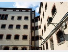 Außenansicht eines historischen Gefängnisgebäudes mit gelblichen Fassaden, Ziegelsteinrahmungen um die vergitterten Fenster und Überwachungskameras in einem engen Innenhof. Der Himmel ist teilweise sichtbar.