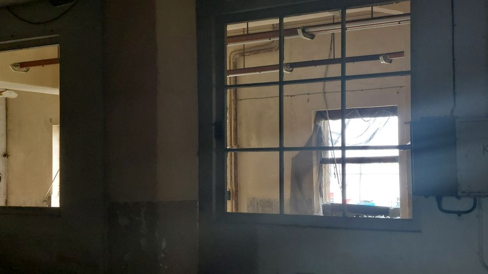 Fenster in einem verlassenen, industriellen Gebäude, mit einer einfachen Metallrahmung und Blick auf eine weitere Wand mit Rohren und einem teilweise verhängten Fenster. Der Raum wirkt durch den Lichteinfall aus den Fenstern hell, aber verlassen.