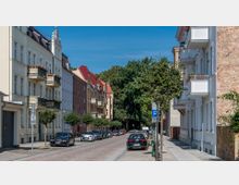 Wohnstraße mit gepflegten, mehrstöckigen Altbauten, teils mit Balkonen, auf beiden Seiten. Die Straße ist mit Kopfsteinpflaster belegt, gesäumt von kleinen Bäumen, und rechts sind geparkte Autos sowie ein Parkplatzschild sichtbar. Im Hintergrund endet die Straße in einer Baumgruppe.